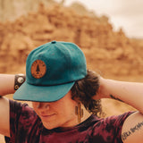 Person in the desert wearing a blue cap and wooden cactus earrrings.