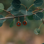 Pair of wooden circular dangle earrings with a green/blue stone in the center hanging from a stick with leaves.