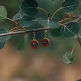 Pair of wooden circular dangle earrings with a green/blue stone in the center hanging from a stick with leaves.