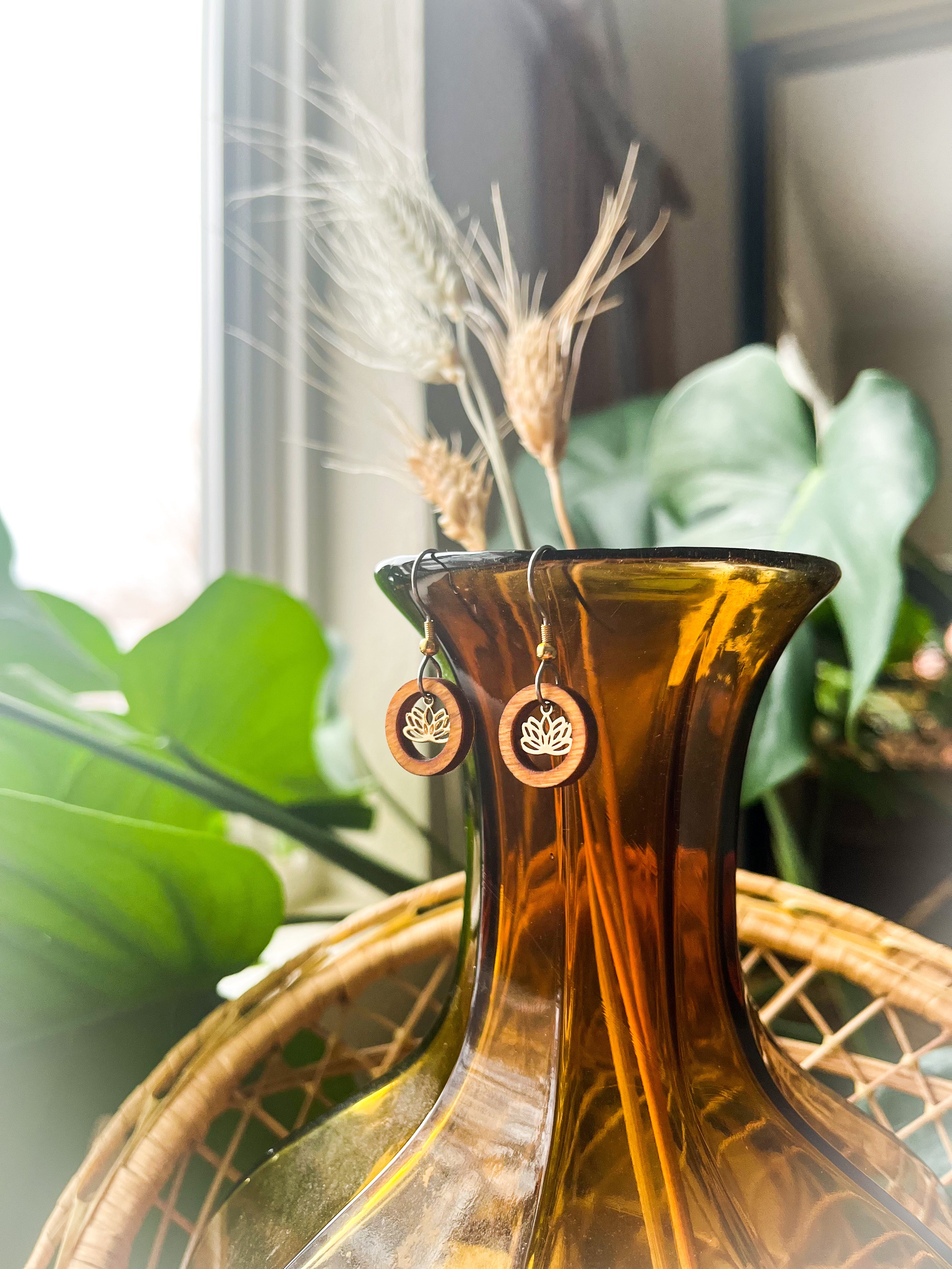 A pair of wooden earrings with a wood ring surrounding a small lotus charm hanging from the top of an orange glass vase.