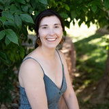 Smiling woman sitting under a tree wearing wooden earrings in the shape of butterfly wings.