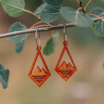 A pair of wooden earrings in the shape of a diamond with a mountain peak carved into the center hanging from a stick with leaves.