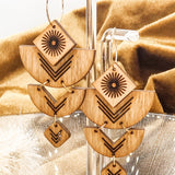Wooden earrings with geometric designs on a stand in front of golden fabric.