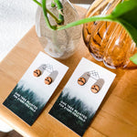 Wooden stud earrings with a mountain etched into them on cards with tree planting message on a wooden surface with vases and plants.