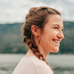 Smiling woman wearing large wooden hoop earring, with a golden ring hanging in the hoop, against a mountain background.