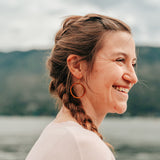 Smiling woman wearing large wooden hoop earring, with a golden ring hanging in the hoop, against a mountain background.