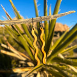 A pair of wooden earrings in a squiggle pattern hanging from a stick in front of a cactus.
