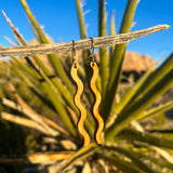 A pair of wooden earrings in a squiggle pattern hanging from a stick in front of a cactus.