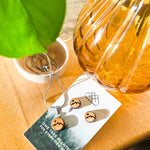 Wooden stud earrings with a mountain peak etched into them on a card with 'lovelevel' branding catching the sun underneath a leaf.